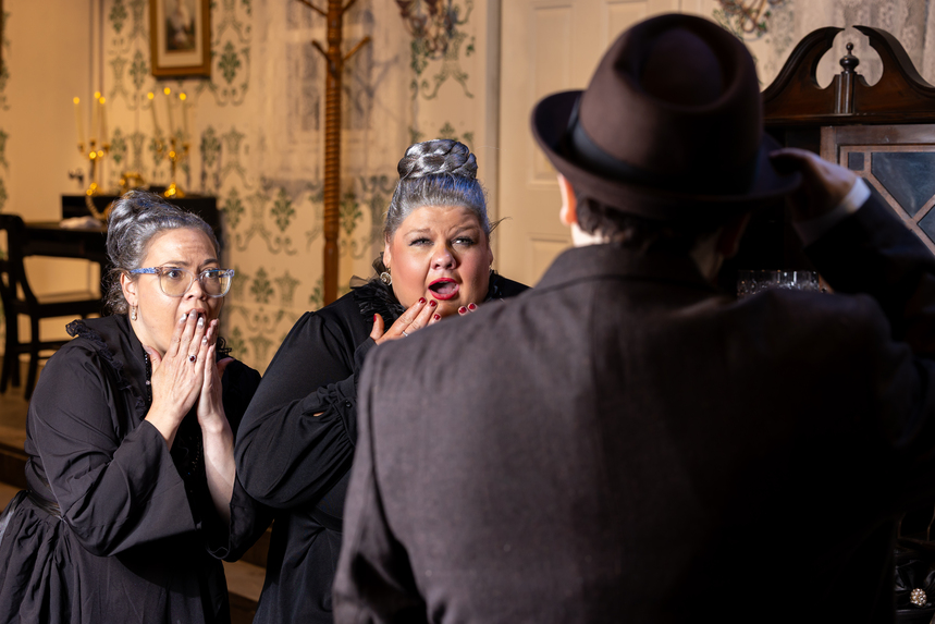 A man in a hat stands with his back to the camera, facing two women in vintage attire, set in a charmingly decorated room.
