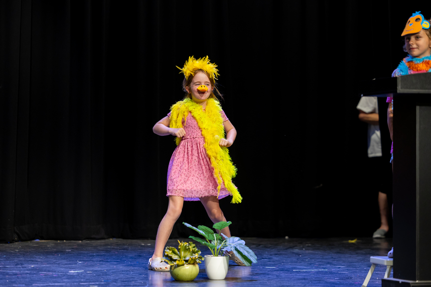 A child in a pink dress and bright yellow feather accessories performs on stage, with decorative plants nearby.