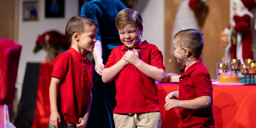 Three children in red polo shirts stand close together, engaged in conversation on stage with festive decorations in the background.